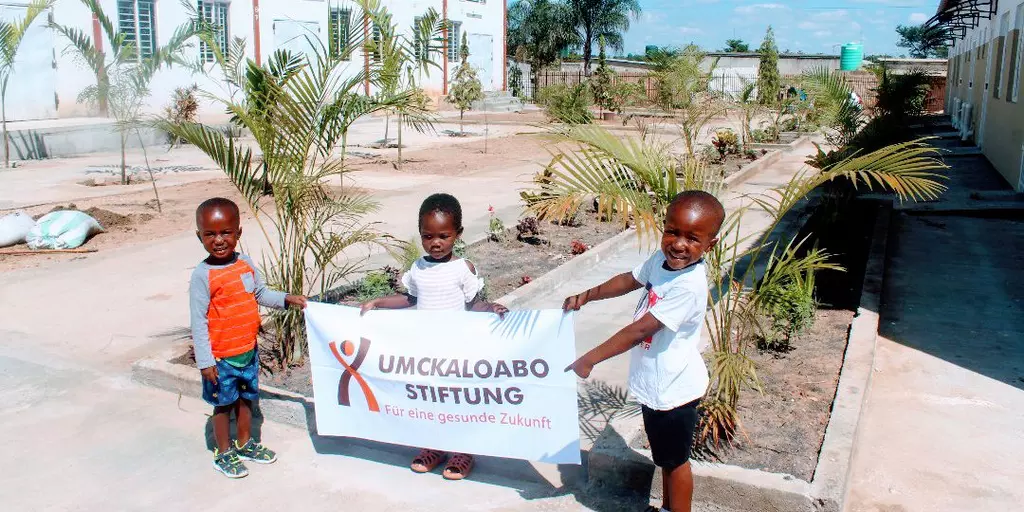 Drei Kinder an der St. Francis-Vorschule halten das Banner der Umckaloabo-Stiftung hoch.