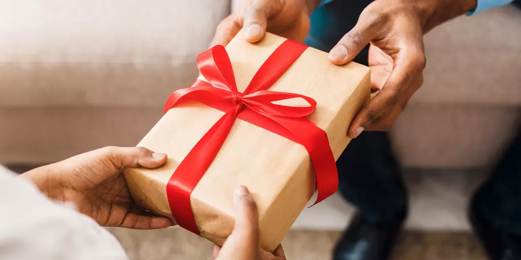Nahaufnahme von zwei Händen, die ein in braunes Papier eingewickeltes Geschenk mit roter Schleife überreichen.