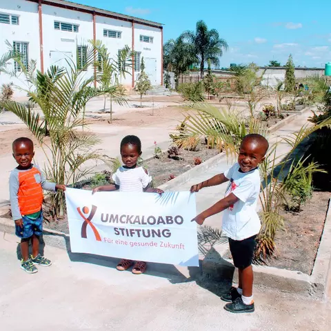 Drei Kinder an der St. Francis-Vorschule halten das Banner der Umckaloabo-Stiftung hoch.