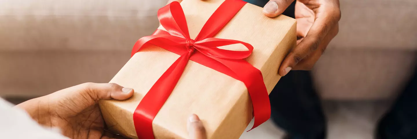 Nahaufnahme von zwei Händen, die ein in braunes Papier eingewickeltes Geschenk mit roter Schleife überreichen.